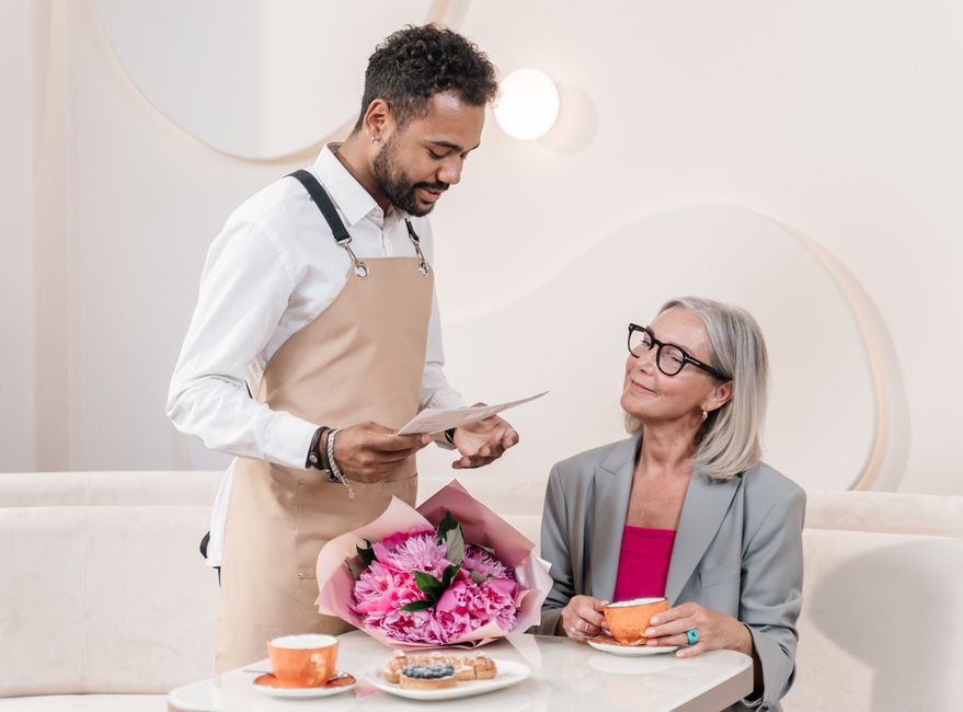 A florist from floristblackwall.co.uk presenting a large bouquet of fresh pink peonies wrapped in soft pink paper, arranged with green foliage, to an elderly woman with gray hair and glasses, who is s