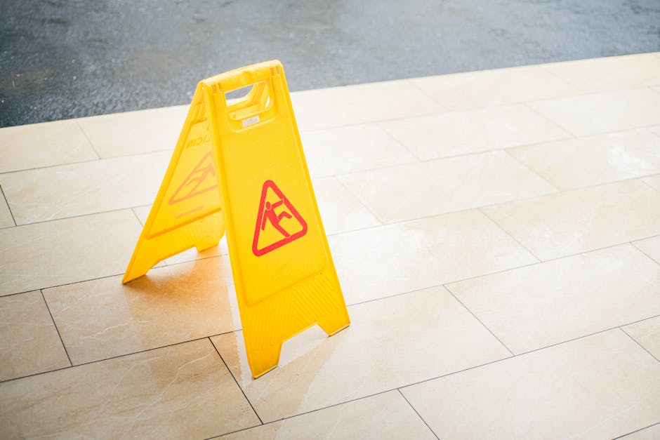 A yellow caution wet floor sign with red warning symbols, positioned on beige tiled flooring near a slightly wet surface, indicating a slip hazard. The sign stands upright, made of plastic, with the w