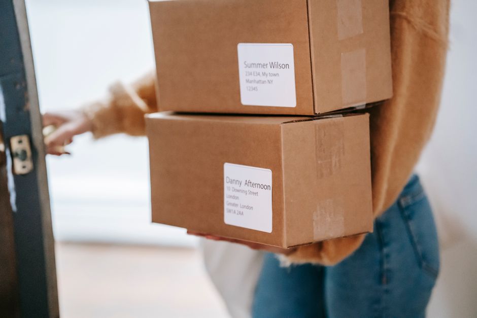 A person wearing a tan sweater and blue jeans is holding two cardboard boxes with white address labels, standing near a door with their hand reaching towards the latch. The boxes are plain brown, seal