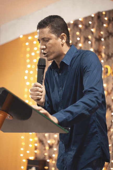 A male florist with short dark hair, wearing a navy blue shirt, is arranging a floral bouquet on a table. The arrangement features an assortment of fresh flowers in shades of pink, purple, and white, 