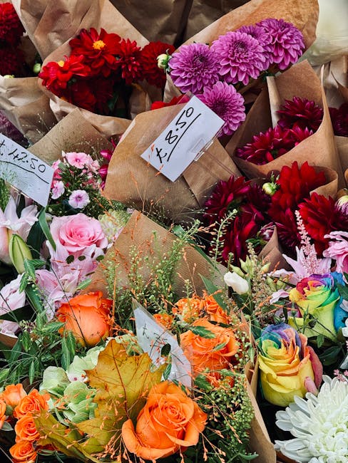 A vibrant assortment of freshly cut flowers arranged in small paper wraps and bouquets at a flower shop, featuring red dahlias, purple chrysanthemums, pink roses, orange spray roses, and multicolored 
