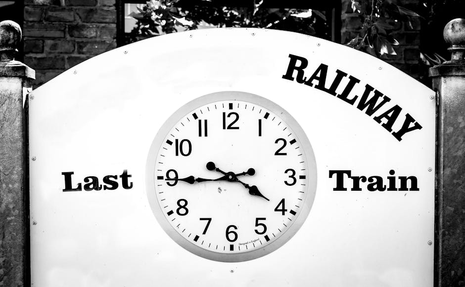 Black and white photograph of a railway station clock, mounted on a white background with a rounded top. The clock face displays black hour, minute, and second hands, showing the time as approximately