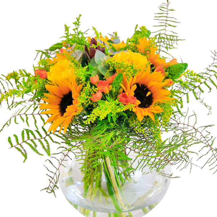 Evoking the lively blend of heritage and modernity that defines Blackwall, this bouquet is a masterful creation balancing classic botanicals with contemporary freshness. At its heart are bold sunflowers, their radiant yellow petals and deep brown eyes conveying optimism and vitality-qualities synonymous with the local community. Surrounding the sunflowers are lemon-yellow garden roses, their petals tightly nested in elegant spirals, symbolizing refinement and joy. Red Hypericum berries are artfully scattered throughout the foliage, their glossy sheen adding a playful accent and evoking memories of autumn festivals by the Thames. Lush greenery abounds: cascading ferns and slender spikes accompany light sprigs of solidago, their tiny golden blooms echoing local park wildflowers. Delicate bluish-green eucalyptus leaves impart both rustic charm and a faint, invigorating scent to the ensemble. Transparent and spherical, the glass vase reveals the arrangement's inner architecture-fresh stems set in crystal water, amplifying the bouquet's sense of clarity and new beginnings. Sunlight streaming over the composition creates a warm, lively ambiance, perfect for celebrations or simple everyday cheer in Blackwall's riverside homes. This arrangement is designed for meaningful moments: birthdays, returns, or tokens of appreciation, crafted with both skill and local pride.